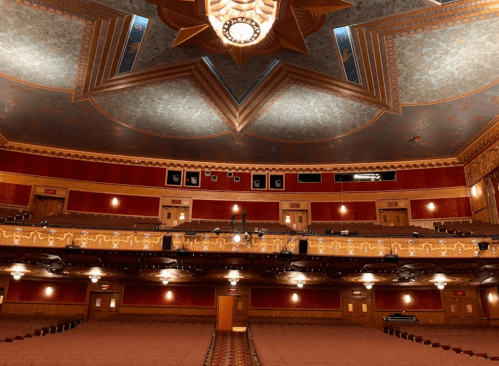 Interior of a grand theater featuring ornate ceiling, red walls, and empty seating, viewed from the stage.