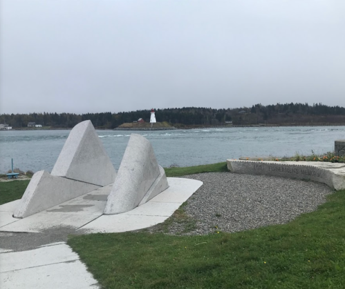 A gray stone memorial with abstract shapes overlooks a calm sea and a distant lighthouse on a cloudy day.
