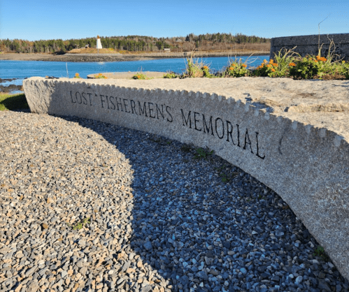 A stone memorial inscribed "Lost Fishermen's Memorial" by a rocky shore with a lighthouse in the background.