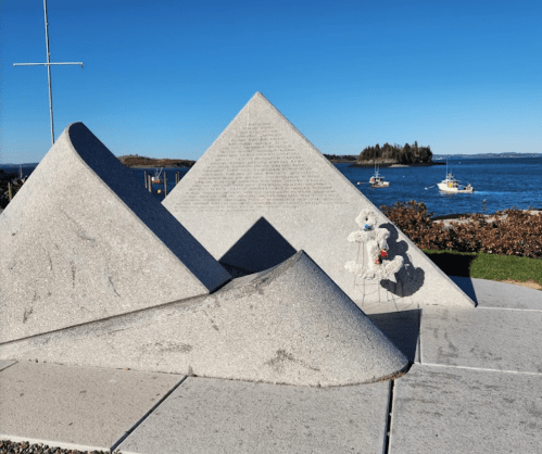 A triangular stone monument by the water, with a small stuffed animal placed on it, against a clear blue sky.