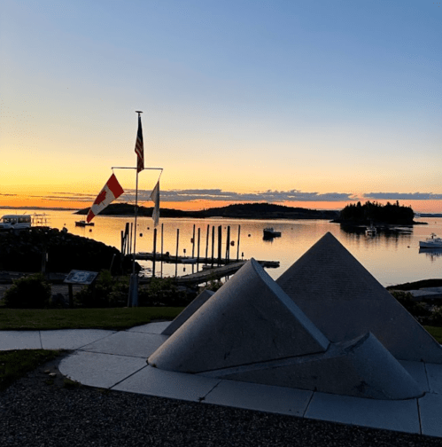 Sunset over a calm harbor with boats, flags, and a geometric sculpture in the foreground.