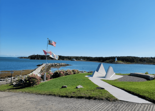 A scenic view of a waterfront with flags, a lighthouse in the distance, and landscaped greenery under a clear blue sky.