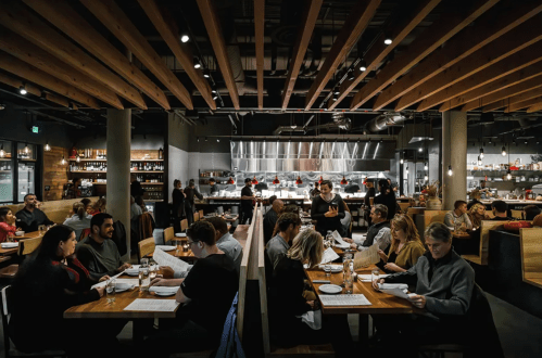 A bustling restaurant interior with diners at wooden tables and an open kitchen in the background. Warm lighting and a lively atmosphere.