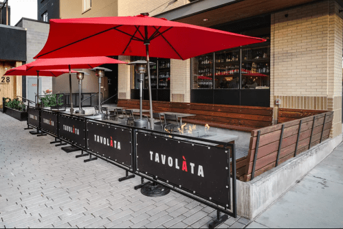 Outdoor dining area with red umbrellas and heaters, featuring a wooden bench and a sign for "Tavolàta."