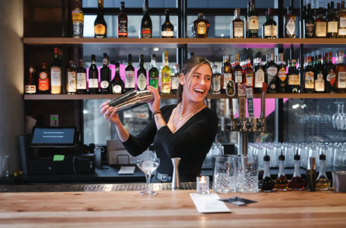 A bartender smiles while shaking a cocktail behind a well-stocked bar with various bottles and glasses.