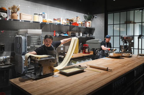 Two chefs in a modern kitchen, one rolling out pasta dough while the other prepares ingredients at a wooden countertop.