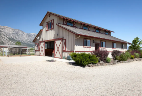A spacious two-story barn-style building with a gravel driveway, surrounded by mountains and greenery.