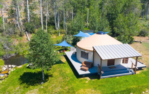 A round yurt with a wooden deck, surrounded by trees and a small stream, set in a green landscape.