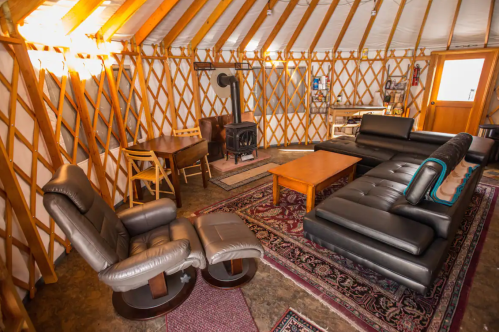 Cozy yurt interior with a black leather sofa, wooden table, chairs, and a wood stove, featuring warm lighting and rustic decor.
