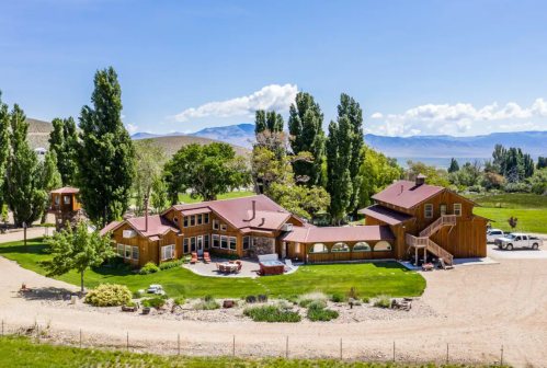 Aerial view of a large wooden house surrounded by greenery and mountains under a clear blue sky.
