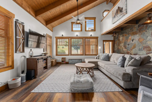 Cozy living room with wooden beams, large windows, a sectional sofa, and a stone accent wall.