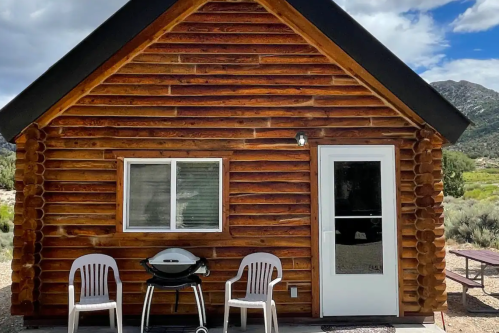 A rustic log cabin with a white door and window, two white chairs, and a grill in front, set against a scenic backdrop.