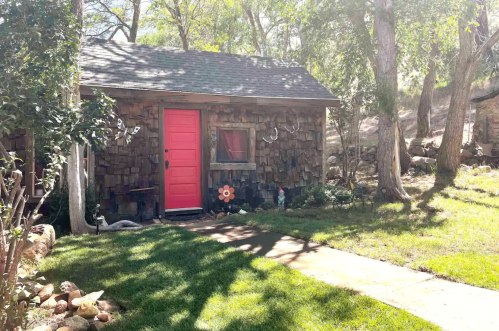 A small rustic cabin with a bright pink door, surrounded by trees and a grassy area.
