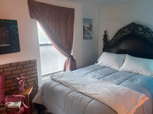 Cozy bedroom featuring a large bed, decorative pillows, a window with curtains, and a vintage armchair.