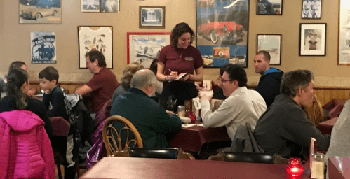 A busy restaurant scene with diners at tables and a waitress taking orders, surrounded by vintage car memorabilia on the walls.