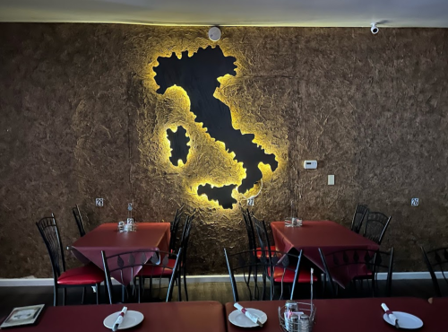 Interior of a restaurant featuring a backlit map of Italy on the wall, with red tables and black chairs.