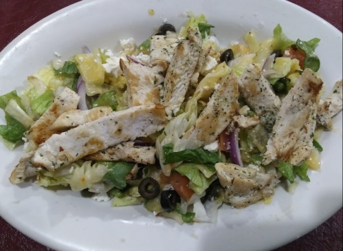 A fresh salad topped with grilled chicken, lettuce, tomatoes, olives, and feta cheese on a white plate.