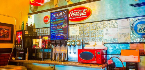 A vintage diner counter with soda dispensers, a Coca-Cola sign, and a menu of daily specials on the wall.