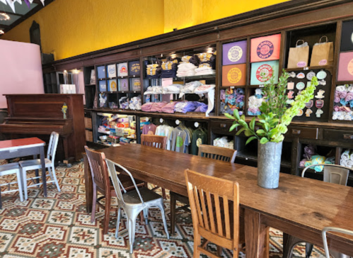 A cozy shop interior featuring a wooden table, chairs, and shelves filled with colorful merchandise against a yellow wall.