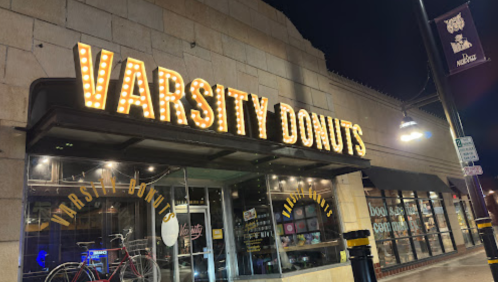 A brightly lit storefront of Varsity Donuts at night, featuring a bicycle parked outside.