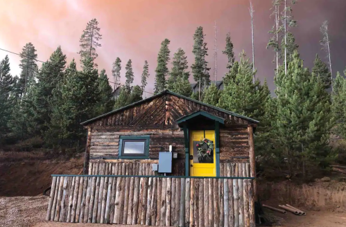 A rustic log cabin surrounded by tall trees, with a yellow door and a wreath, against a smoky, orange sky.
