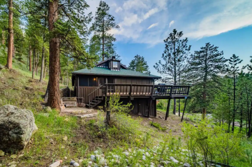 A modern cabin nestled among tall trees, featuring a deck and surrounded by lush greenery and rocky terrain.