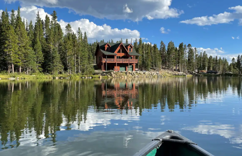 A serene lake scene with a wooden house surrounded by trees, reflecting in the calm water under a blue sky.