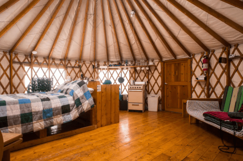 Interior of a cozy yurt featuring a bed, wooden furniture, a kitchenette, and wooden floors. Natural light filters in.