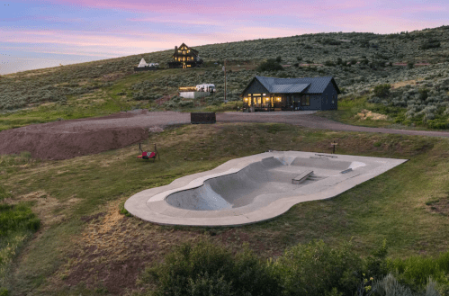 A skate bowl in a grassy area with a house and hills in the background under a colorful sunset sky.