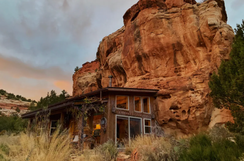 A rustic wooden cabin nestled against a large rock formation, surrounded by sparse vegetation and a colorful sky.