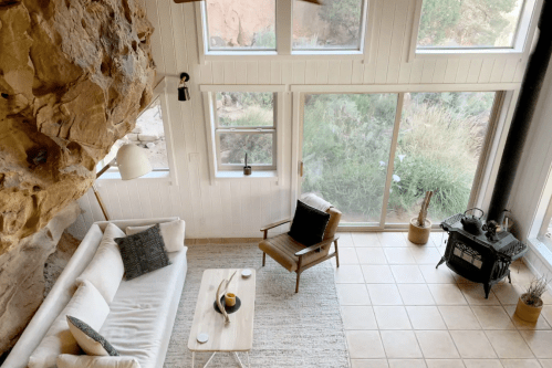 Cozy living space with a rock wall, large windows, a sofa, wooden chair, and a small table, surrounded by greenery.