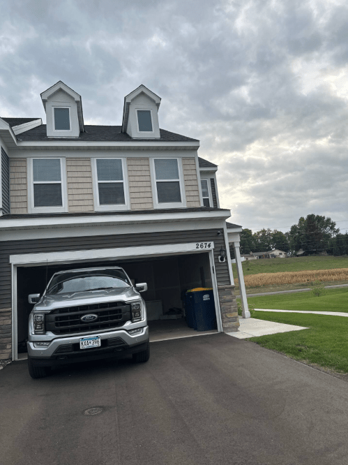 A gray truck parked in a driveway of a two-story house with a garage, under a cloudy sky.