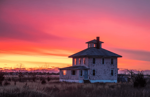A weathered house stands in a field against a vibrant sunset sky with shades of pink, orange, and purple.
