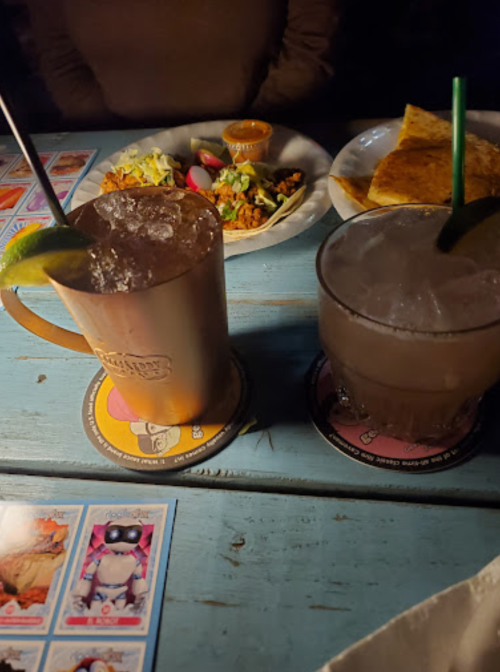 Two cocktails on a table with food in the background, surrounded by colorful coasters and dim lighting.