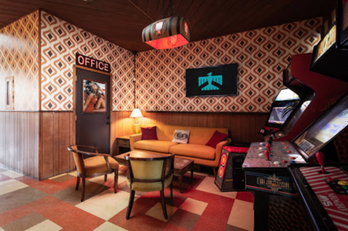 Retro-style lounge with orange couch, patterned wallpaper, and arcade machines in a cozy corner.