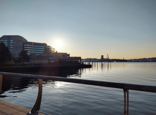 Sunrise over a calm waterfront, with buildings and a pier reflected in the water.