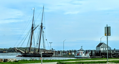 A tall ship docked at a harbor with a small boat nearby and a building labeled "Fisher" in the background.