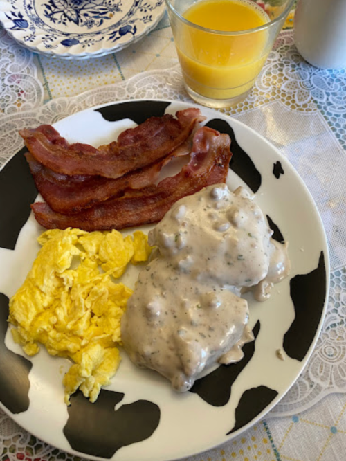 A plate with scrambled eggs, bacon, and biscuits topped with sausage gravy, alongside a glass of orange juice.