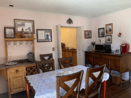 A cozy dining area with a wooden table, lace tablecloth, and vintage decor on the walls, leading to a kitchen.