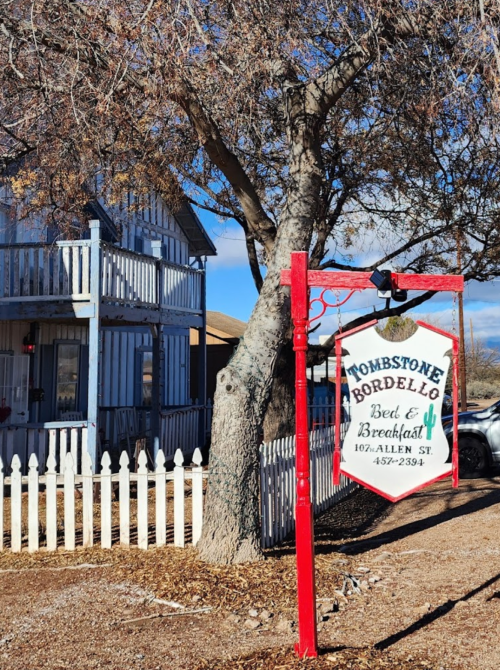 Sign for "Tombstone Bordello Bed & Breakfast" in front of a historic building with a tree and white picket fence.