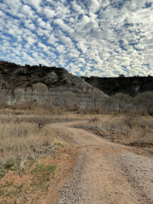 A winding dirt path leads through a dry landscape with sparse vegetation and a cloudy sky above.