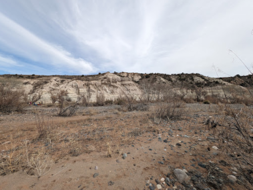 A barren landscape with dry, rocky ground and a distant, layered hillside under a cloudy sky.