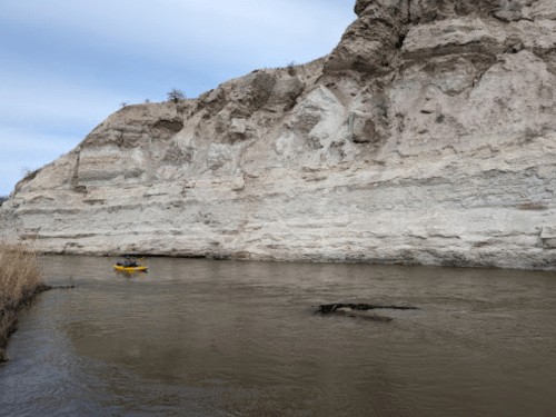 A kayaker paddles on a river beside a steep, rocky cliff under a cloudy sky.