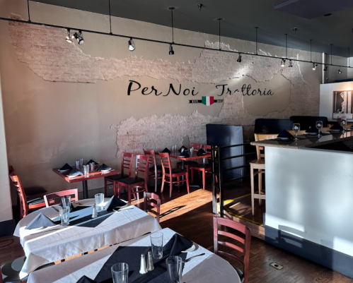 Interior of Per Noi Trattoria, featuring wooden tables, chairs, and a rustic brick wall with the restaurant's name.