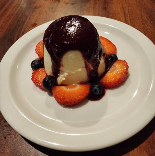 A creamy dessert topped with dark sauce, surrounded by fresh strawberries and blueberries on a white plate.