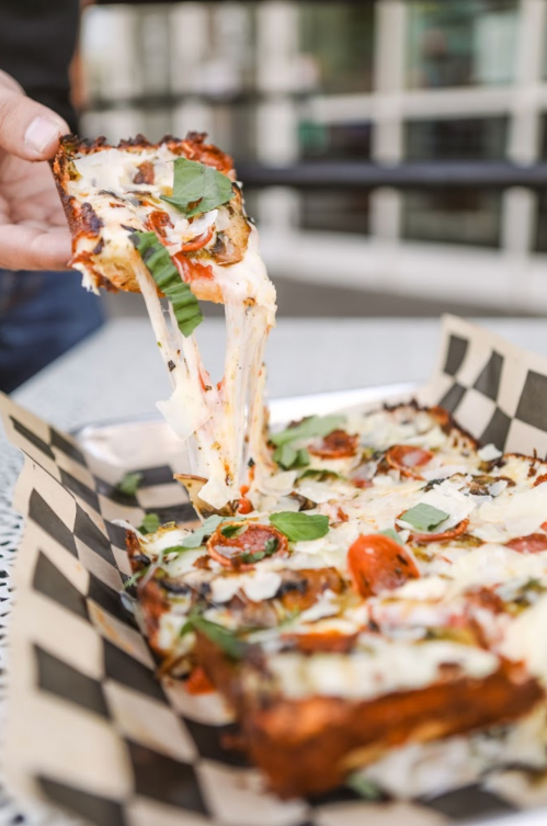 A hand lifts a slice of cheesy pizza topped with tomatoes and basil from a tray on a checkered paper.