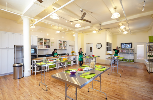 A bright kitchen with three people preparing food at stainless steel tables, surrounded by shelves and modern appliances.