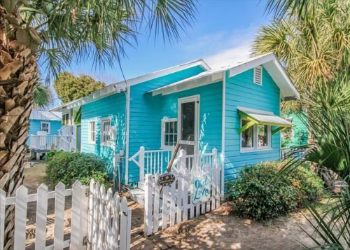 Bright blue cottage with white trim and a picket fence, surrounded by palm trees and sunny skies.