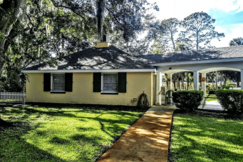 A charming yellow house with green shutters, surrounded by lush grass and trees, featuring a pathway leading to the entrance.