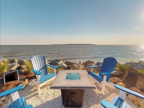 A serene beach scene with blue chairs around a fire pit, overlooking calm waters and a distant shoreline.